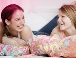 Bilde av to unge kvinner som ligger i en seng og ler. Bildet illustrerer at i puberteten skjer mange endringer i kroppen, men det er helt normalt.
